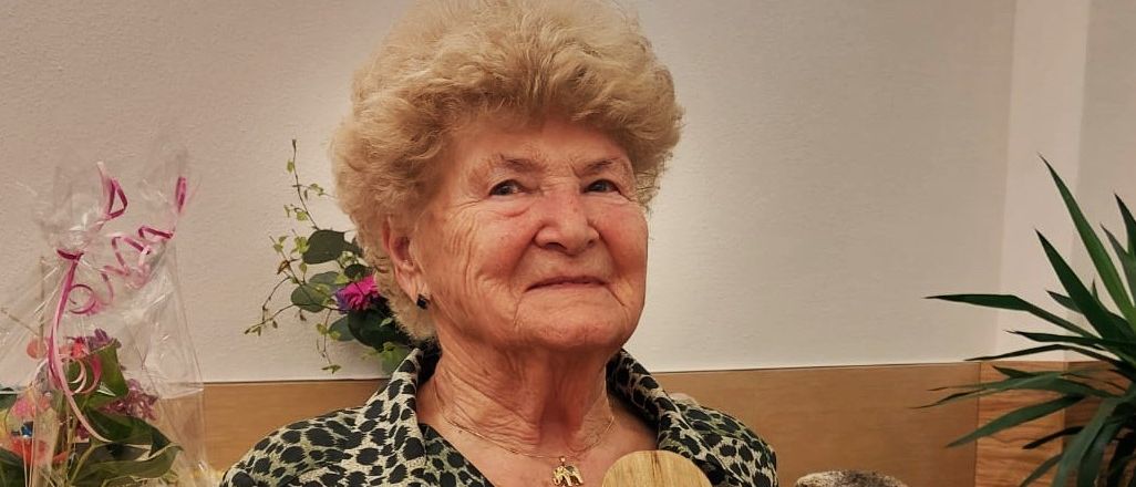 Eine ältere Frau hält eine Holztafel mit gravierten Rosen und Text, sitzt drinnen vor einem Schreibtisch mit Pflanzen und Blumen.