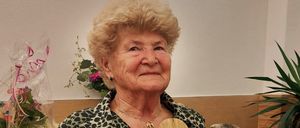 Eine ältere Frau hält eine Holztafel mit gravierten Rosen und Text, sitzt drinnen vor einem Schreibtisch mit Pflanzen und Blumen.