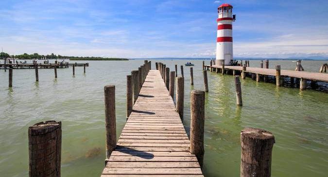 Ein langer Holzsteg ragt ins Wasser und endet mit einem Leuchtturm auf der rechten Seite. Das Wasser ist grün, und der Himmel ist blau mit einigen Wolken.