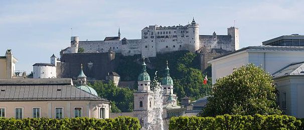 Ein Blick auf einen Park mit einem Brunnen, Hecken und einer Burg auf einem Hügel. Bäume, Büsche und Menschen sind im Bereich verstreut.