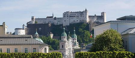 Ein Blick auf einen Park mit einem Brunnen, Hecken und einer Burg auf einem Hügel. Bäume, Büsche und Menschen sind im Bereich verstreut.
