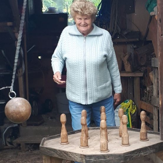 Eine ältere Frau in einem blauen Pullover steht in einer Werkstatt und lächelt. Vor ihr steht ein Holzspiel mit neun Holzkegeln.