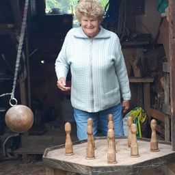 Eine ältere Frau in einem blauen Pullover steht in einer Werkstatt und lächelt. Vor ihr steht ein Holzspiel mit neun Holzkegeln.