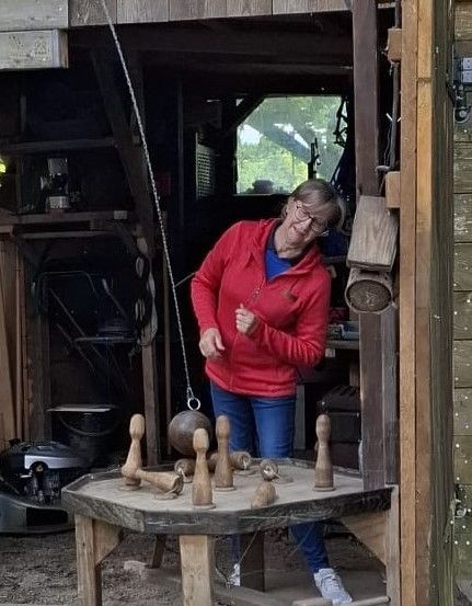 Eine Frau in einer roten Jacke steht in einem Holzschuppen und spielt ein Spiel mit Holzstiften und Bällen auf einem Tisch.