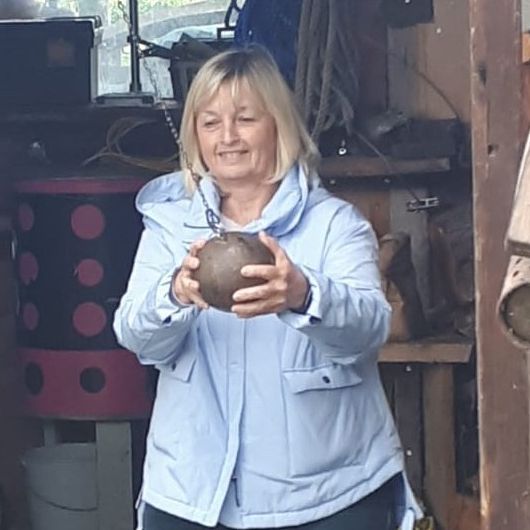 Eine Frau in einer blauen Jacke steht in einem Schuppen, hält einen braunen Ball und hat vor sich hölzerne Objekte und einen Holztisch.