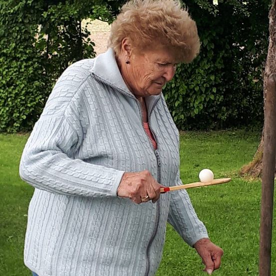 Eine ältere Frau in einem hellblauen Pullover spielt draußen Tischtennis und hält einen Holzschläger mit einem weißen Ball.