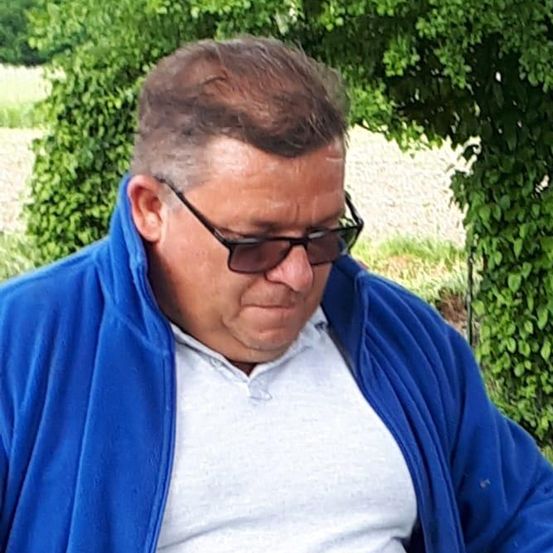 Ein Mann in einem blauen Jacke hält einen Holzstab mit einem weißen Ball darauf und steht auf einem grünen Rasen.