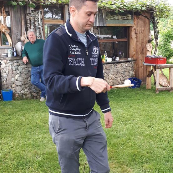 Ein Mann in einer blauen Jacke mit dem Wort Yacht wirft einen Ball auf einem Rasen. Ein anderer Mann in Grün steht neben einer Steinmauer. Dahinter befindet sich ein Holzgebäude.