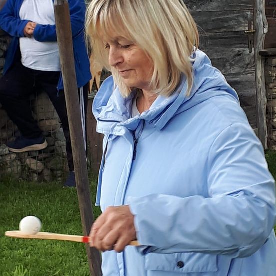 Eine Frau in einer blauen Jacke hält einen Holzstab mit einem Ball und übt auf einer Grasfläche. Hinter ihr steht eine weitere Person in der Nähe eines Holzzauns.