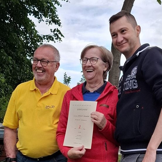 Drei Personen stehen auf einem Rasenfeld. Die Frau in der Mitte hält ein Zertifikat. Der Mann links trägt ein gelbes Poloshirt. Der Mann rechts trägt ein schwarzes Sweatshirt.