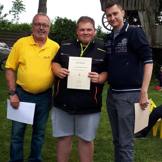 Drei Männer stehen auf einer Wiese und halten Papiere in den Händen. Der Mann in der Mitte hält ein Zertifikat mit der Nummer 3. Der Mann auf der linken Seite trägt ein gelbes Hemd und eine Brille. Der Mann auf der rechten Seite trägt einen dunkelblauen Mantel.