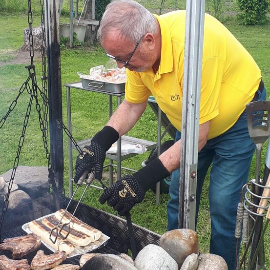 Ein Mann in einem gelben Shirt und schwarzen Handschuhen kocht Fleisch auf einem Grill im Freien. Der Grill steht auf einer Steinfläche mit Steinen und Felsen.