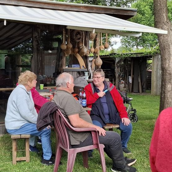 Drei Erwachsene sitzen auf Stühlen in einem schattigen Bereich im Freien. Eine Frau hält einen Stift und schaut auf ein Tablet, während zwei andere in ein Gespräch vertieft sind. Flaschen und Becher stehen auf dem Tisch, und mehrere Gegenstände hängen von der Decke.