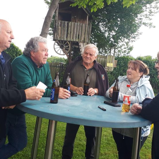 Fünf Erwachsene versammeln sich um einen Tisch in einem Park, mit Flaschen und einem Handy auf dem Tisch, ein Baumhaus im Hintergrund.