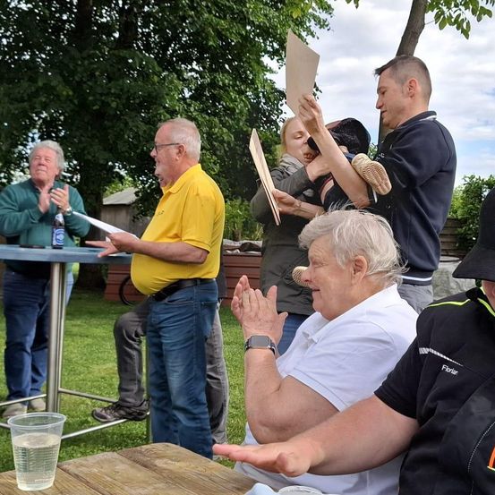 Eine Gruppe von Menschen versammelt sich im Freien, möglicherweise auf einer Party, und jubelt und klatscht. Ein Mann hält ein Papier, während andere ebenfalls Papiere halten. Ein Tisch mit einem Becher und einer Flasche befindet sich in der Nähe.