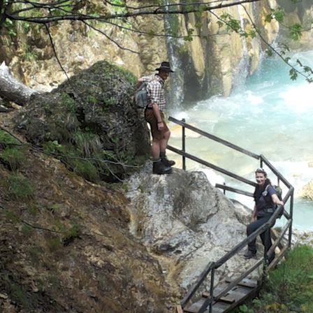 Zwei Wanderer klettern einen steilen Felsen mit einem Metallgeländer hinauf, einer trägt einen Hut und der andere hat einen Rucksack. Der Wasserfall fließt die Felswand hinunter.