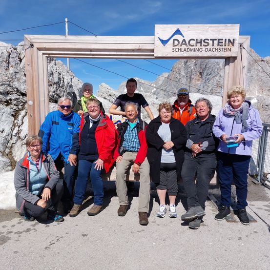 Eine Gruppe von Menschen posiert für ein Foto vor dem Dachstein Schädling-Dachstein-Schild vor einem Berg.