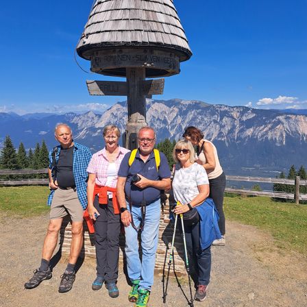 Fünf Wanderer posieren für ein Foto vor einem Bergwegweiser mit der Aufschrift 'En Romanen Slaven Ger'.