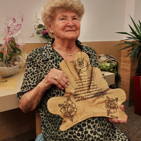 Eine ältere Frau sitzt in einem Raum mit einem Holzbrett mit einer Rose und Text. Sie trägt ein Leoparden-Kleid und einen Ring. Ein Blumenvase steht auf dem Schreibtisch.