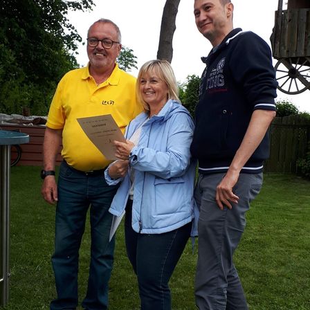 Drei Personen stehen in einem Garten; eine Frau hält ein Zertifikat und lächelt. Ein Mann in einem gelben Hemd und ein anderer in einem dunkelblauen Sweatshirt begleiten sie. Im Hintergrund befinden sich ein Holzrad und ein Zaun.