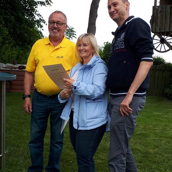 Drei Personen stehen in einem Garten; eine Frau hält ein Zertifikat und lächelt. Ein Mann in einem gelben Hemd und ein anderer in einem dunkelblauen Sweatshirt begleiten sie. Im Hintergrund befinden sich ein Holzrad und ein Zaun.