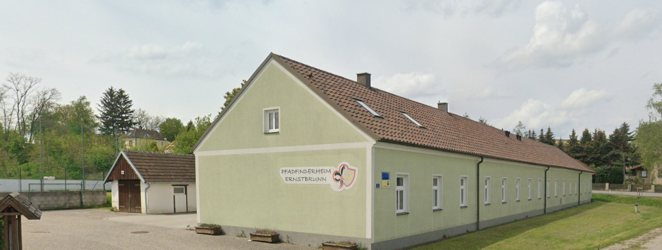 Ein beigefarbenes Gebäude mit einem braunen Dach, weißen Fenstern und einem Logo an der Wand, mit einem kleineren Gebäude dahinter und einem Grasbereich davor.