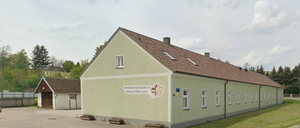 Ein beigefarbenes Gebäude mit einem braunen Dach, weißen Fenstern und einem Logo an der Wand, mit einem kleineren Gebäude dahinter und einem Grasbereich davor.