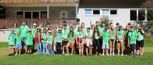 Eine Gruppe junger Menschen in grünen Shirts und Shorts steht vor einem Haus. Sie lächeln und scheinen für ein Foto zu posieren.