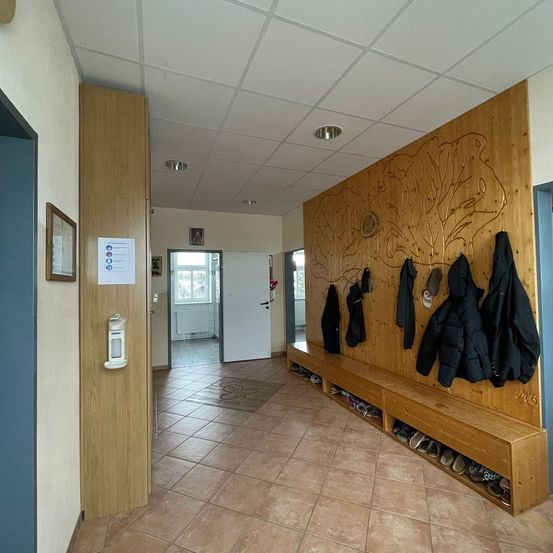 Ein Flur mit Holzböden, Wänden und einer Decke. Eine Holzbank mit Haken hält Jacken und Schuhe. Eine offene weiße Tür führt in einen Raum mit Fenster.