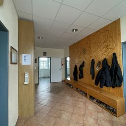 Ein Flur mit Holzböden, Wänden und einer Decke. Eine Holzbank mit Haken hält Jacken und Schuhe. Eine offene weiße Tür führt in einen Raum mit Fenster.