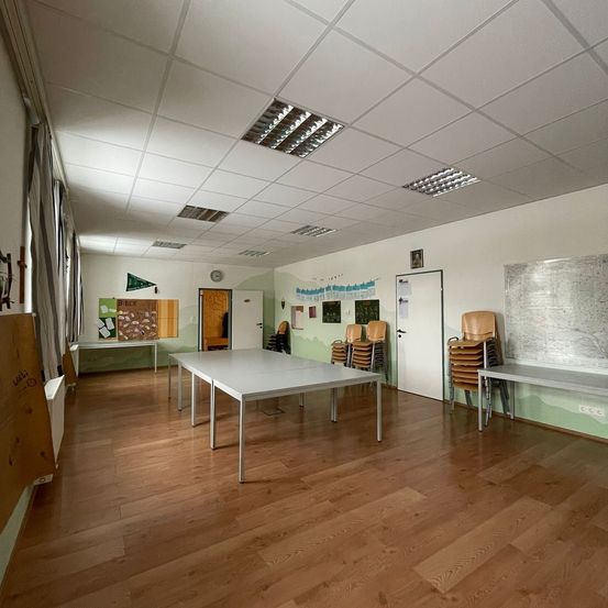 Ein leerer Raum mit Holzboden, weißen Wänden und einer Decke mit quadratischen Lichtern. Zwei Tische und Stühle sind rechts gestapelt. Eine Anschlagtafel und eine Uhr befinden sich links.