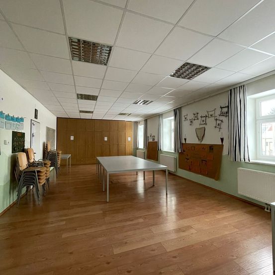 Ein leerer Raum mit Holzböden, einem weißen Tisch und einer Reihe gestapelter Stühle. Die Wände sind grün, und es gibt einen Holzschrank mit mehreren Türen. Ein Fenster mit einem Heizkörper darunter und eine Anschlagtafel mit verschiedenen Papieren und Zeichnungen befindet sich an der Wand.