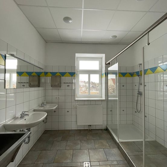 Ein Badezimmer mit weißen Fliesen, zwei Waschbecken, einem Heizkörper, einer Glasdusche und einem Fenster. Die Wände haben blaue und gelbe Verzierungen.