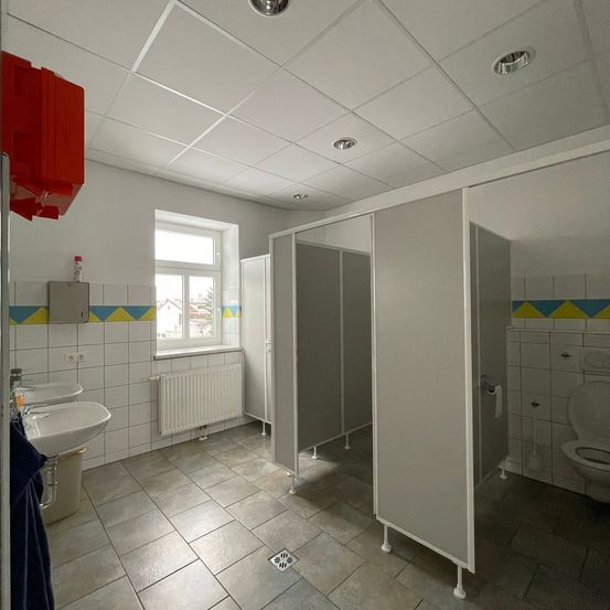 Ein leeres Badezimmer mit weißen Fliesen, drei weißen Kabinen, einem Waschbecken und einem roten Seifenspender. Die Decke ist weiß mit eingelassenen Leuchten.