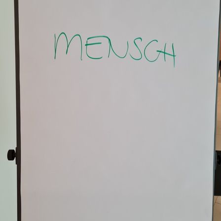Eine Whiteboard mit grünem Text, der 'MENSCH' liest. Das Board ist an einer Wand mit schwarzem Rand montiert. Der Raum scheint gut beleuchtet zu sein.