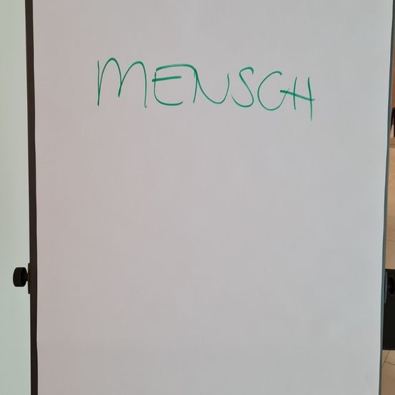 Eine Whiteboard mit grünem Text, der 'MENSCH' liest. Das Board ist an einer Wand mit schwarzem Rand montiert. Der Raum scheint gut beleuchtet zu sein.