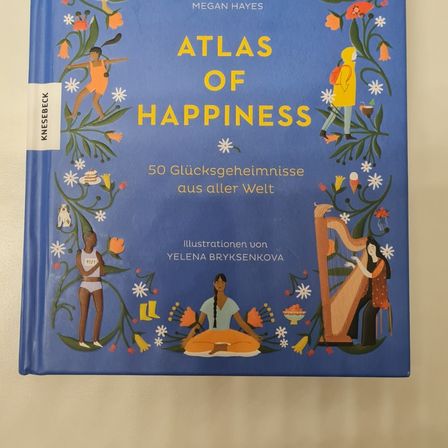 Das Buchcover von Atlas of Happiness von Megan Hayes zeigt farbenfrohe Illustrationen und einen blauen Hintergrund. Es enthält Illustrationen von Menschen aus verschiedenen Kulturen, einer Harfenspielerin und einer Läuferin.