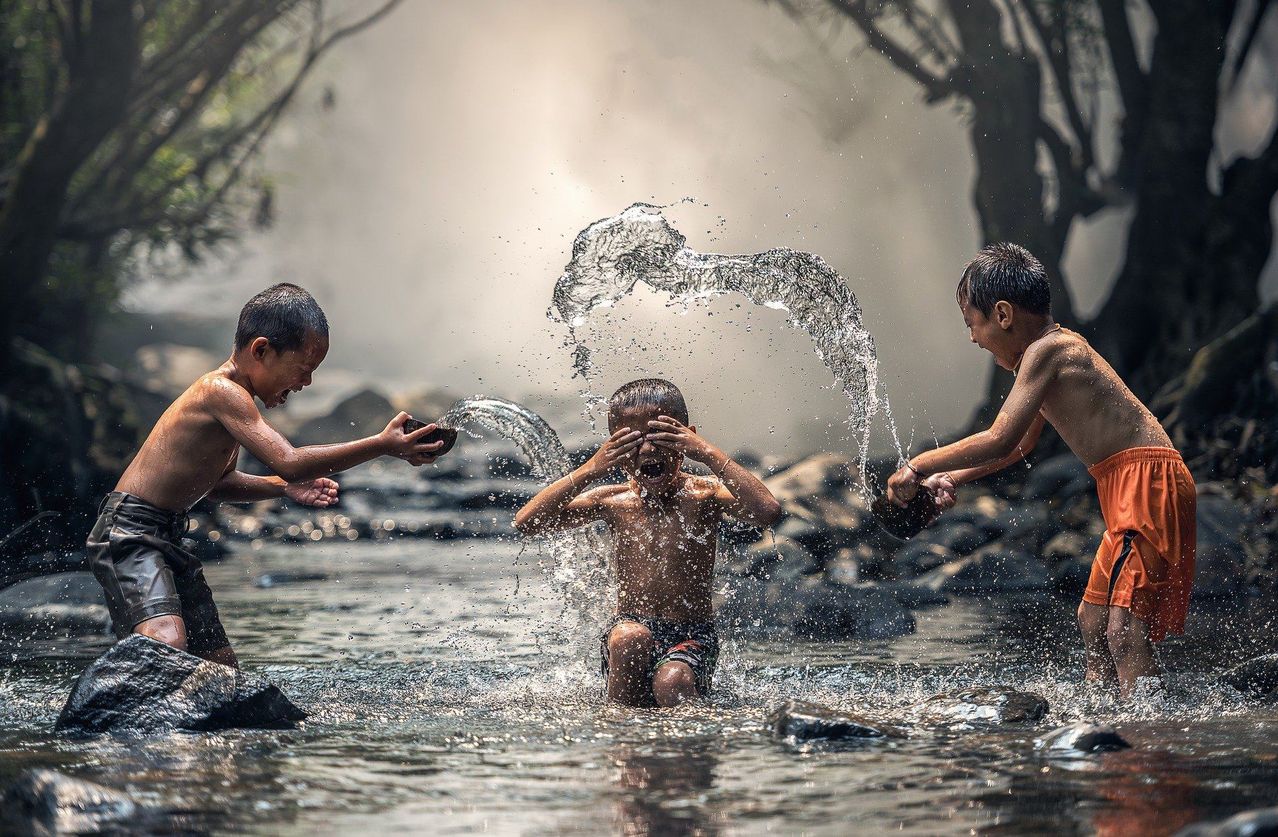 Drei Jungen spielen in einem Fluss, einer hält einen Stein und spritzt Wasser. Ein anderer Junge schützt sein Gesicht vor dem Wasser, während der dritte Junge einen Stein hält.