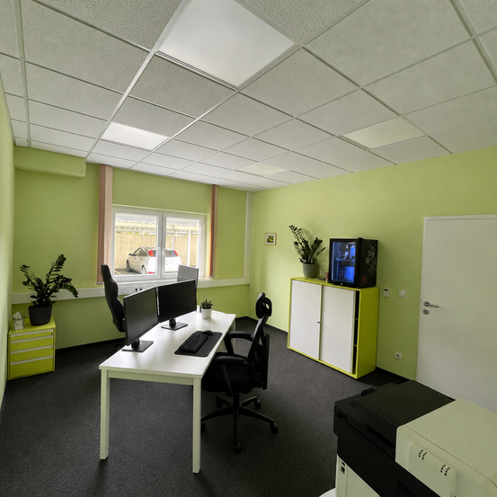 Ein Büro mit einem weißen Schreibtisch und zwei Monitoren. Eine Topfpflanze steht links. Ein gelber Schrank mit einem Fernseher steht rechts. Ein schwarzer Stuhl steht vor dem Schreibtisch. Eine weiße Tür steht rechts.