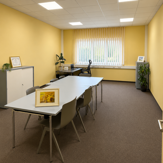 Ein leeres Büro mit gelben Wänden, einem langen Tisch und einem Fenster mit Jalousien. Es gibt Topfpflanzen, gerahmte Bilder und einen Schreibtisch mit Laptop.