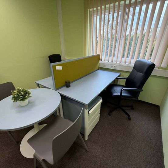 Ein leeres Büro mit einem Schreibtisch, einem runden Tisch und zwei Stühlen. Ein kleiner Topf mit Pflanzen steht auf dem Tisch. Der Raum hat eine grüne Wand, ein Fenster mit Jalousien und einen Teppichboden.