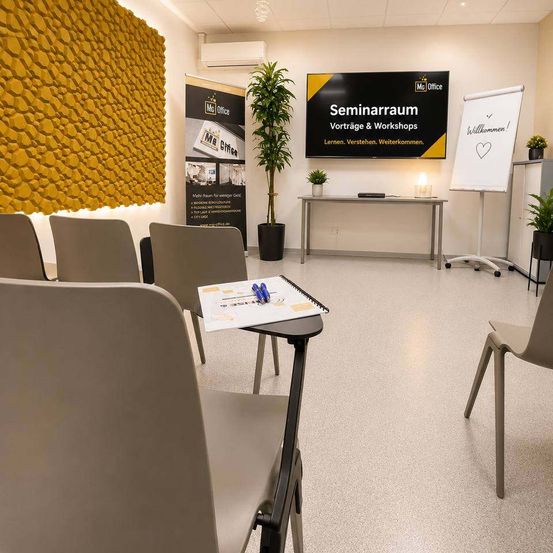 Ein Seminarraum mit Stühlen, einem Schreibtisch, einer Tafel, einem Monitor, einer Lampe und einer Pflanze. Der Raum hat eine gelbe Wand und Klimaanlage.