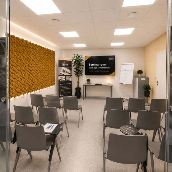 Ein Seminarraum mit in Reihen angeordneten Stühlen, einer gelben strukturierten Wand und einem Tisch mit Pflanzen. Ein Whiteboard und ein schwarzes Schild sind im Raum aufgestellt. Eine Glastür befindet sich auf der linken Seite.