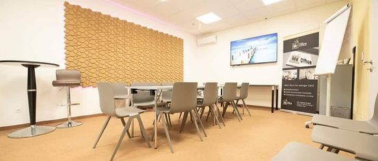 An empty meeting room with beige walls, a patterned wall, and a table with gray chairs. A TV and an advertisement board are mounted on the wall. A gray bar stool is beside the wall.