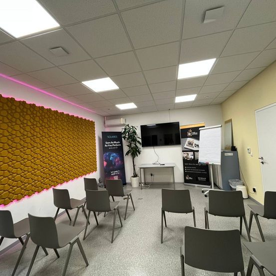 Ein Konferenzraum mit grauen Stühlen, einem Schreibtisch, einem an der Wand montierten Fernseher und einer Whiteboard. Die Wände sind gelb und weisen ein strukturiertes Muster auf.