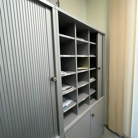 Ein grauer Schrank mit vertikalen Streifen auf den Türen, gefüllt mit ordentlich angeordneten Akten und Dokumenten auf Regalen. Der Raum hat helle Wände und einen Vorhang.