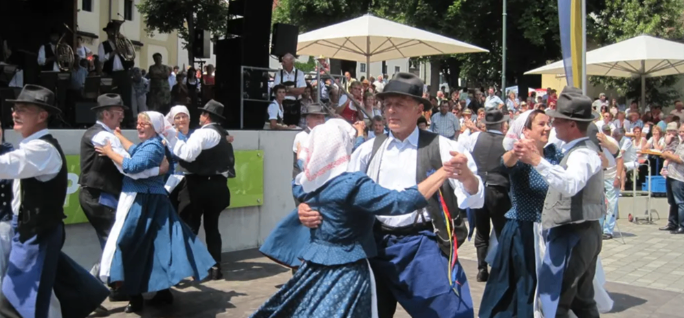 Mehrere Menschen tanzen in traditionellen Kostümen auf einem Betonboden. Sie halten sich an den Händen. Dahinter steht eine Menschenmenge, die zuschaut. Es gibt Bäume und eine Bühne mit einem weißen Schirm und Lautsprechern.