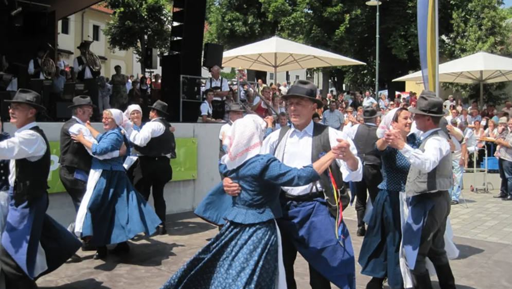 Mehrere Menschen tanzen in traditionellen Kostümen auf einem Betonboden. Sie halten sich an den Händen. Dahinter steht eine Menschenmenge, die zuschaut. Es gibt Bäume und eine Bühne mit einem weißen Schirm und Lautsprechern.