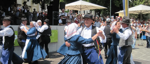 Mehrere Menschen tanzen in traditionellen Kostümen auf einem Betonboden. Sie halten sich an den Händen. Dahinter steht eine Menschenmenge, die zuschaut. Es gibt Bäume und eine Bühne mit einem weißen Schirm und Lautsprechern.