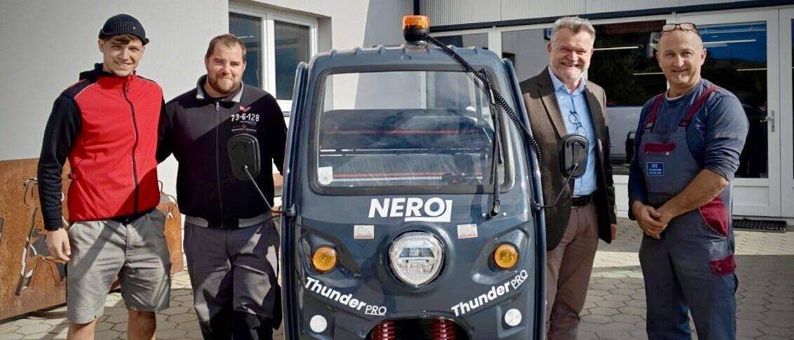 Vier Männer posieren für ein Foto neben einem blauen Fahrzeug mit dem Wort Nero darauf. Der Mann auf der linken Seite trägt eine rote Jacke und Turnschuhe.